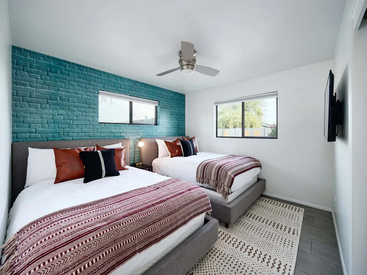 A guest bedroom with two beds adorned with decorative throws, a turquoise brick accent wall, wall-mounted TV, and a stylish ceiling fan. The space is relaxed, comfortable, and ideal for families or friends sharing a room.