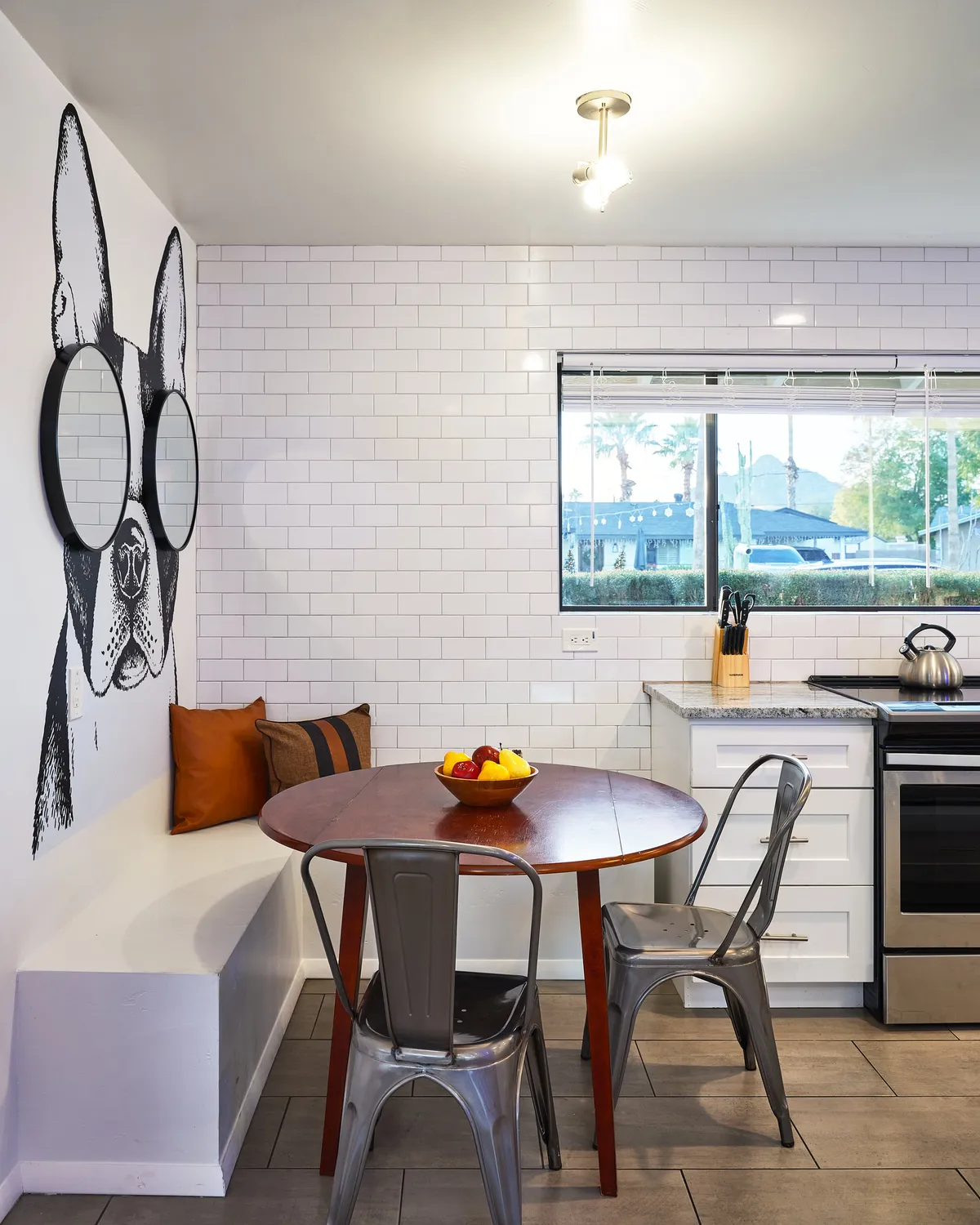 A bright and cheerful breakfast nook anchored by a round wooden table surrounded by silver chairs. The playful Frenchie dog mural on the wall adds a whimsical touch, while light filters in from a nearby kitchen window, creating an inviting spot for morning coffee or brunch.
