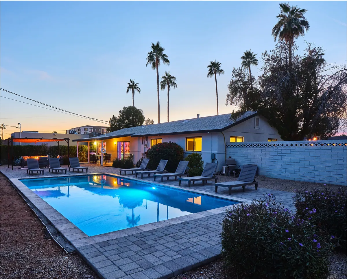The image showcases the home's inviting backyard at golden hour, featuring a sparkling swimming pool framed by lush palm trees and modern lounge chairs. The stylish exterior of the house serves as a backdrop, suggesting plenty of space for relaxation or gatherings. The tranquil setting hints at both daytime sunbathing and evening poolside fun under the desert sky.