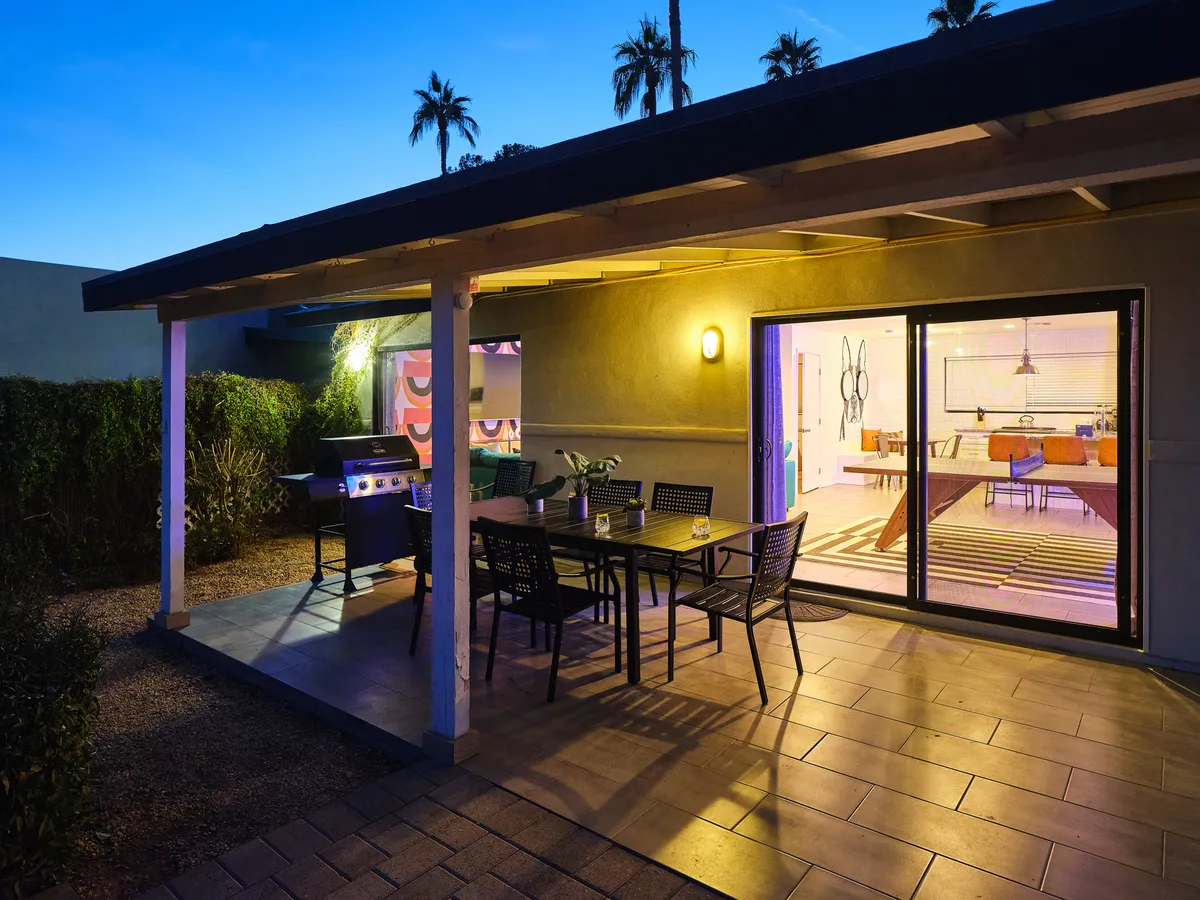 This image features an outdoor dining table and grill positioned right outside a large sliding glass door, visually linking the interior and exterior. The setup is perfect for alfresco meals and lively gatherings, with interior decor visible through the glass and a welcoming sunset glow.