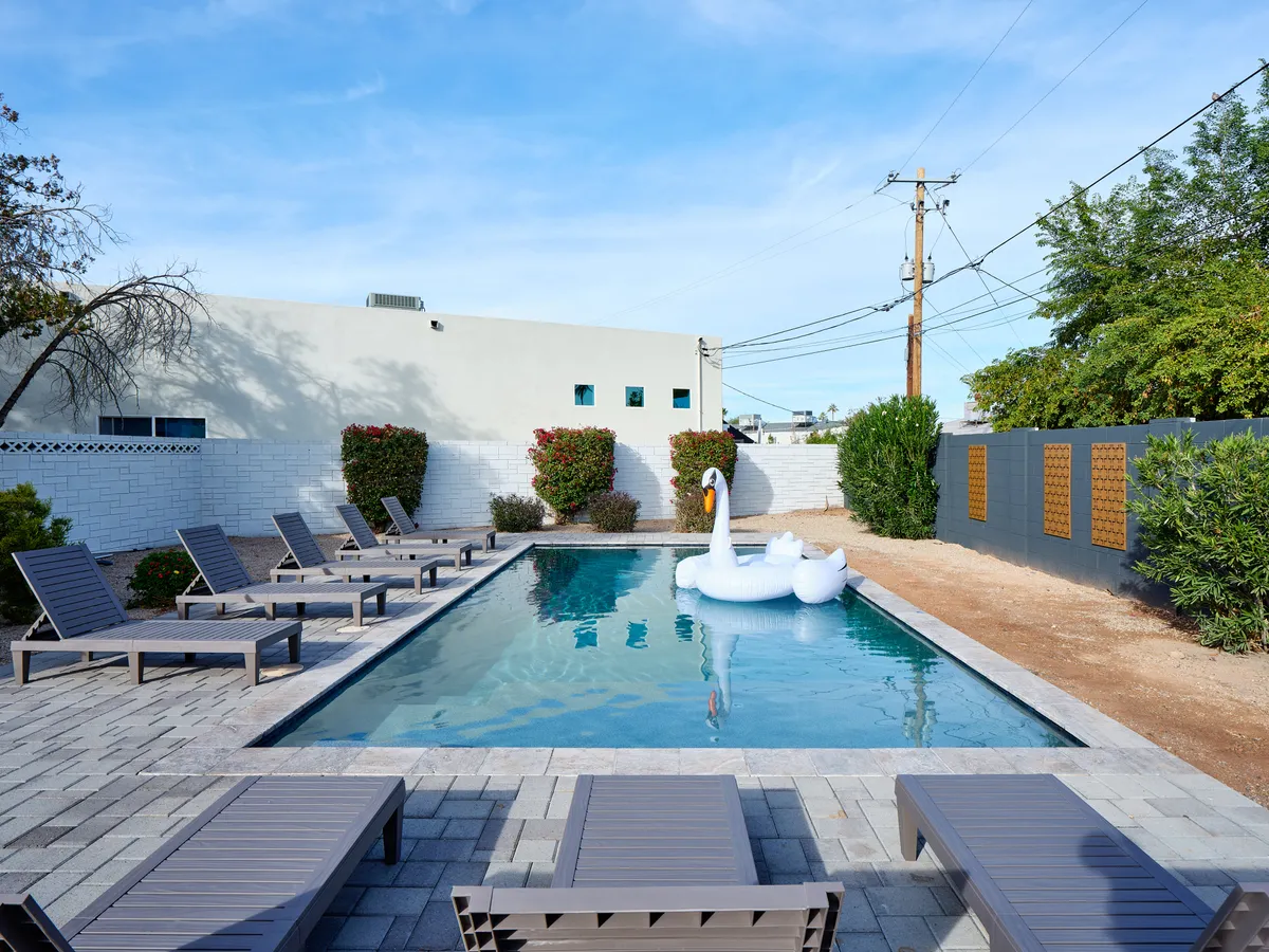 A sparkling backyard swimming pool surrounded by lush landscaping, lounge chairs, and a playful swan float. The scene invites relaxation, family fun, and socializing, with space for both energetic games and peaceful lounging under sunny skies.