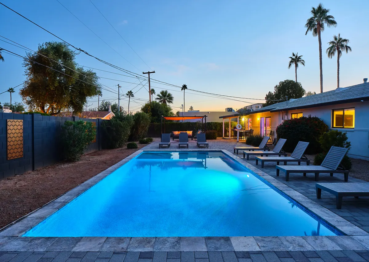 Here, the backyard is displayed with a focus on the inviting swimming pool, comfortable lounge chairs, and an adjacent outdoor seating area shaded by tall palm trees. The mood is leisurely and tranquil, ideal for both sunbathing and group relaxation.