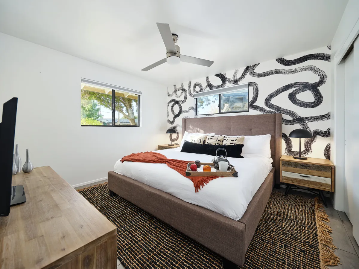 A sunlit bedroom featuring a bed with a decorative throw, artistic wall design, and nightstands with lamps. Natural light pours in through the window, creating a cozy, inviting atmosphere perfect for winding down or enjoying a quiet morning.