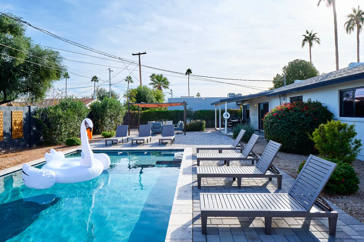 A vibrant backyard scene featuring a sparkling swimming pool with a whimsical swan float, surrounded by stylish lounge chairs, lush landscaping, and swaying palm trees. The sunny setting beckons for leisure, fun in the water, and social gatherings.