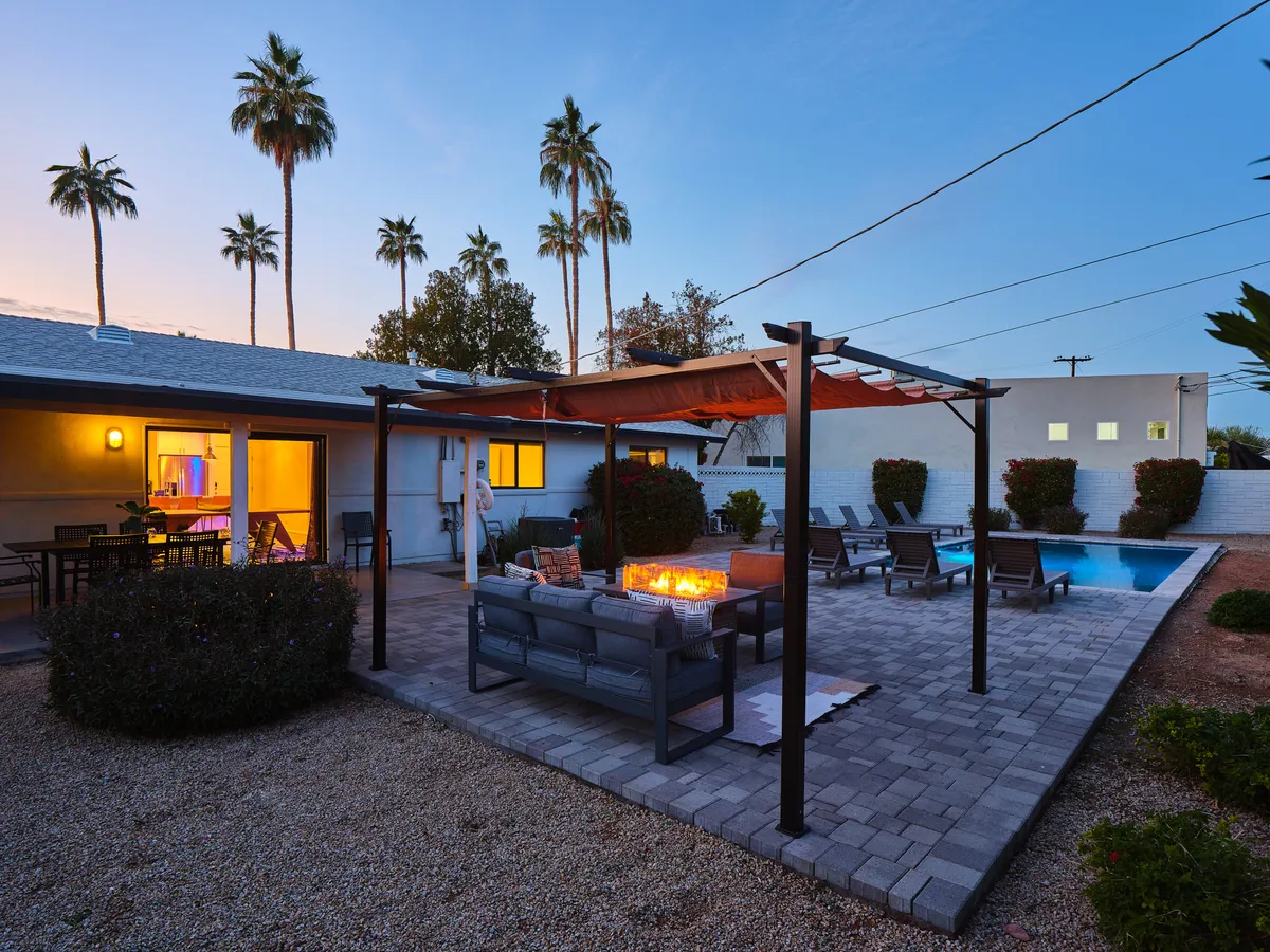 This exterior image features a cozy fire pit surrounded by comfortable outdoor seating, with a swimming pool and palm trees visible in the background. The space is warmly lit, evoking a sense of relaxation and inviting social gatherings.