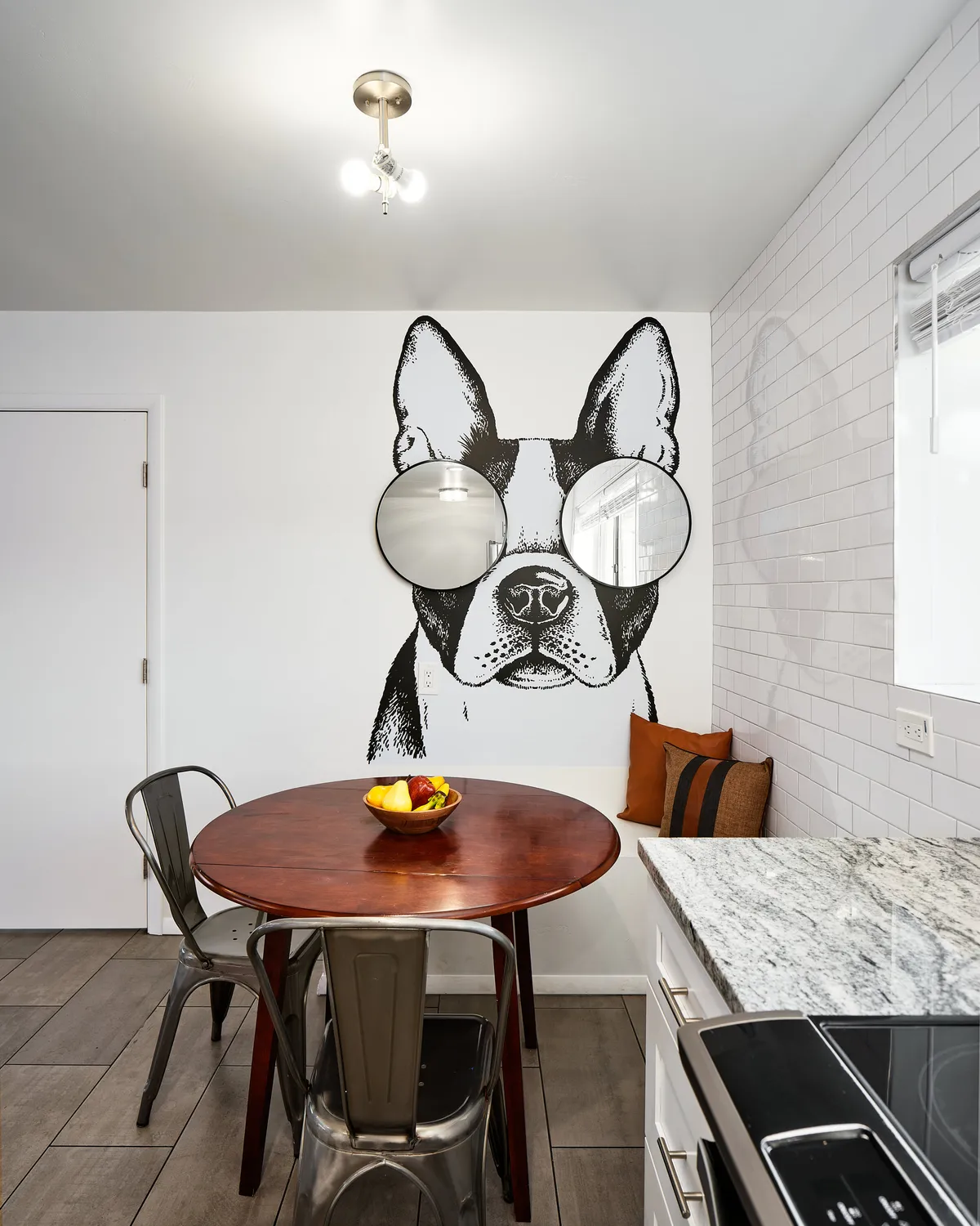 A close-up of the whimsical breakfast nook with a round wooden table, metal chairs, a fruit bowl centerpiece, and the standout Frenchie mural as the backdrop. The scene is staged for casual dining or a cozy brunch, radiating a quirky and inviting charm.
