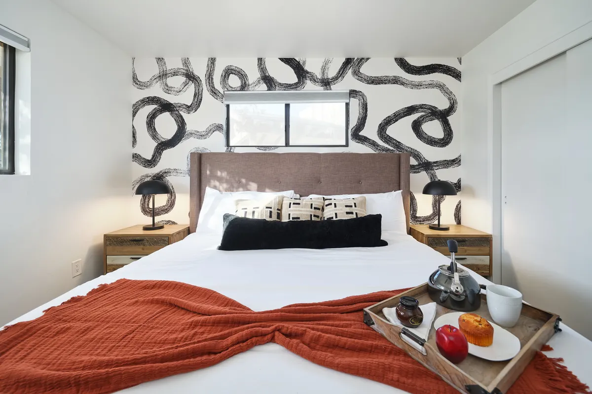 A cozy bedroom scene featuring a bed with decorative pillows, artistic wall design, and a breakfast tray with food and drink. The space feels inviting and personal, perfect for starting the day or enjoying quiet moments.