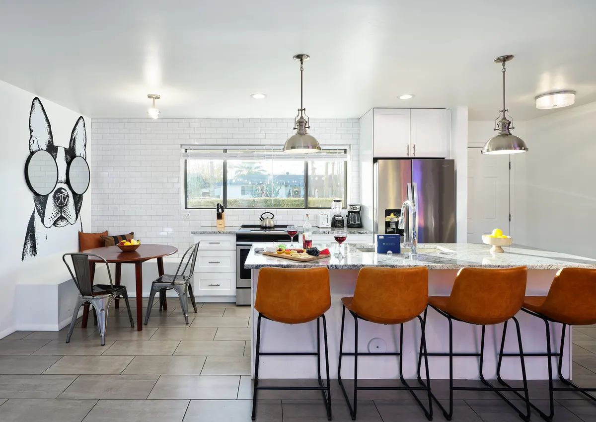 This image captures a bright, open kitchen and dining area featuring a vibrant dog mural on the wall, a kitchen island with bar stools for casual seating, and a dining table set with fruit and drinks. The stainless steel appliances are subtly visible, and the cheerful decor creates an inviting atmosphere ideal for socializing and dining.